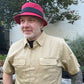 A man wearing a red bucket hat with a black band and a beige short-sleeve button-up shirt, standing outdoors in front of a white brick wall and garden plants.