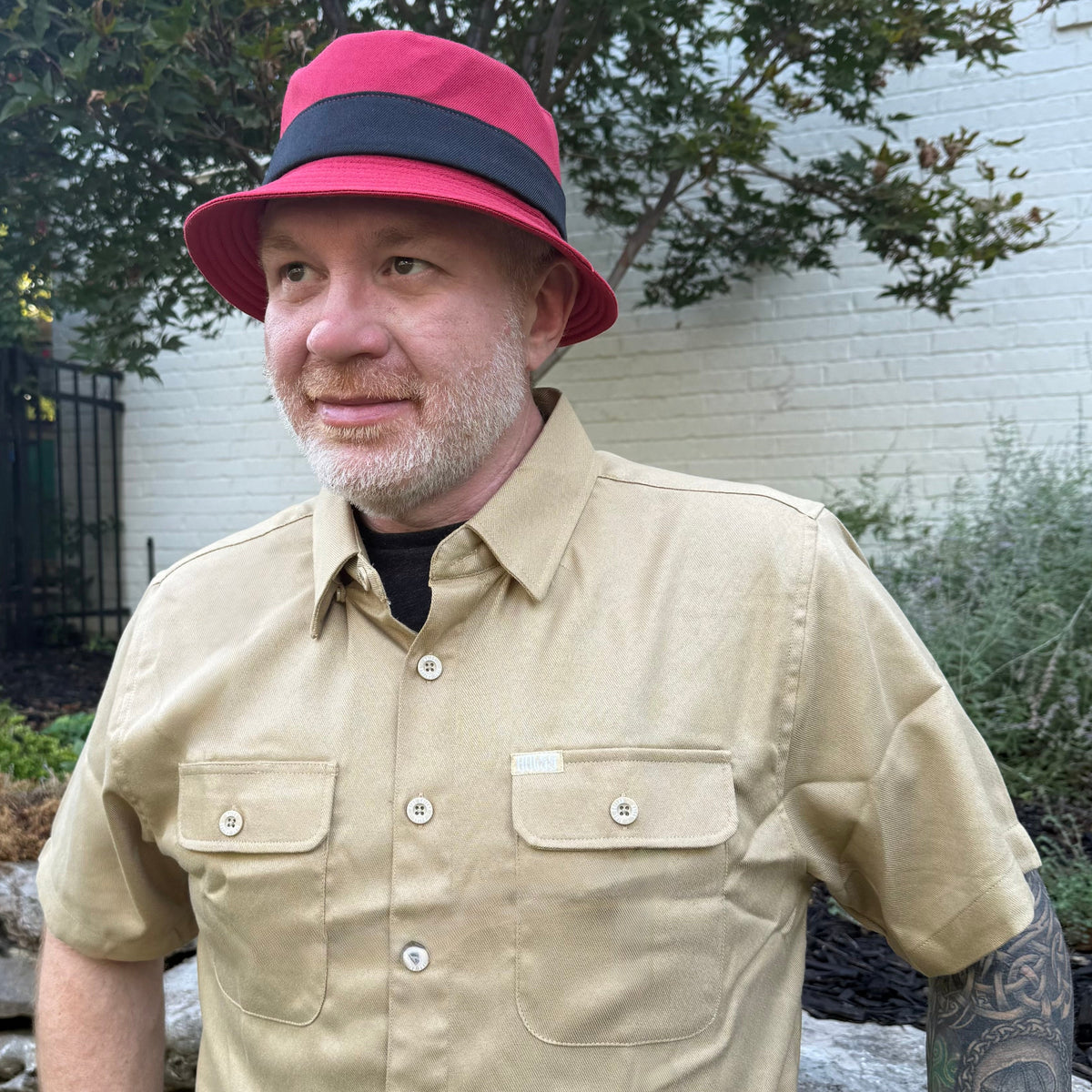 A man wearing a red bucket hat with a black band and a beige short-sleeve button-up shirt, standing outdoors in front of a white brick wall and garden plants.