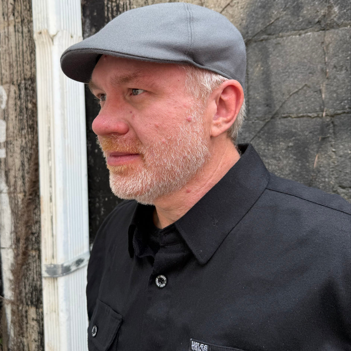 A man wearing a gray flat cap and a black button-up shirt, standing outdoors against a textured gray wall.