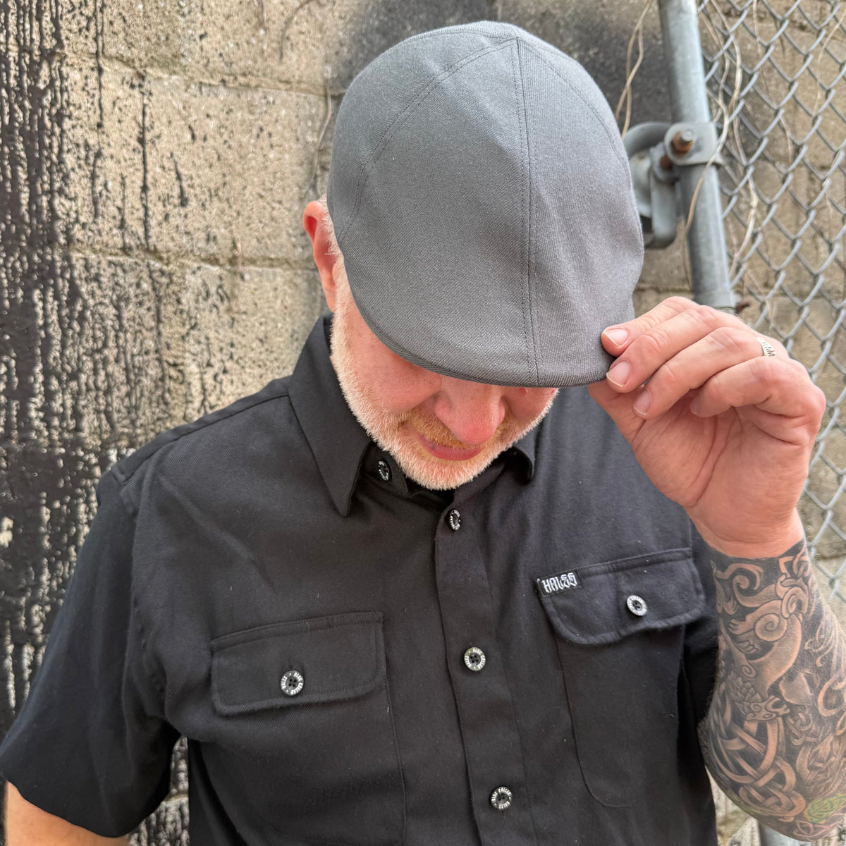 A man wearing a gray flat cap and a black short-sleeve button-up shirt with chest pockets, standing outdoors against a concrete wall and chain-link fence.