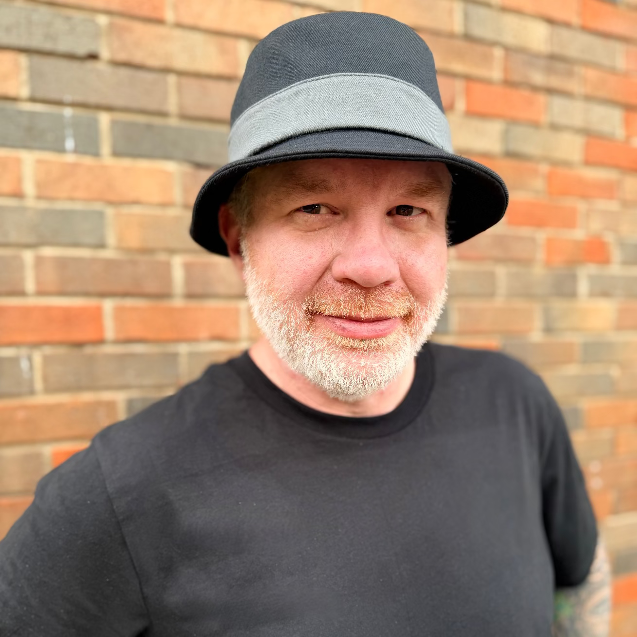 Man wearing a black bucket hat and black shirt against a brick wall.