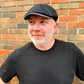Man wearing a black cap and black t-shirt standing in front of a brick wall.