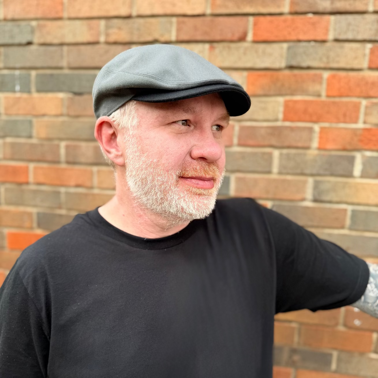 Man wearing a gray cap and black t-shirt standing against a brick wall.