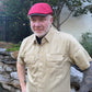A man wearing a beige short-sleeve button-up shirt and a red flat cap, standing outdoors in a backyard setting with trees, a stone wall, and a fence visible.