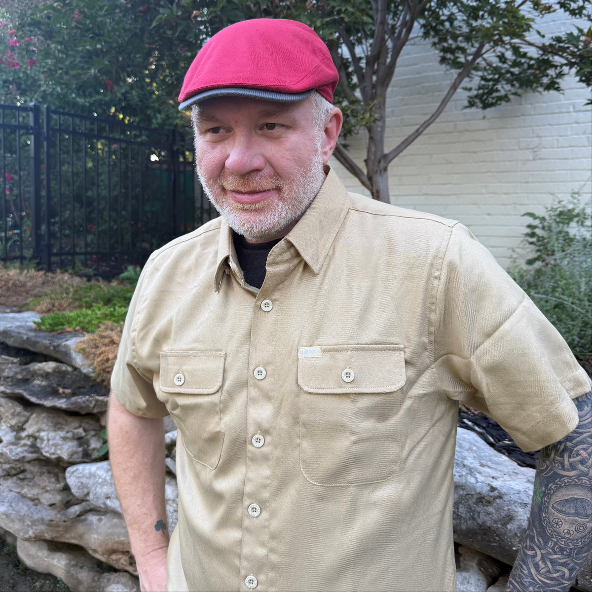 A man wearing a beige short-sleeve button-up shirt and a red flat cap, standing outdoors in a backyard setting with trees, a stone wall, and a fence visible.