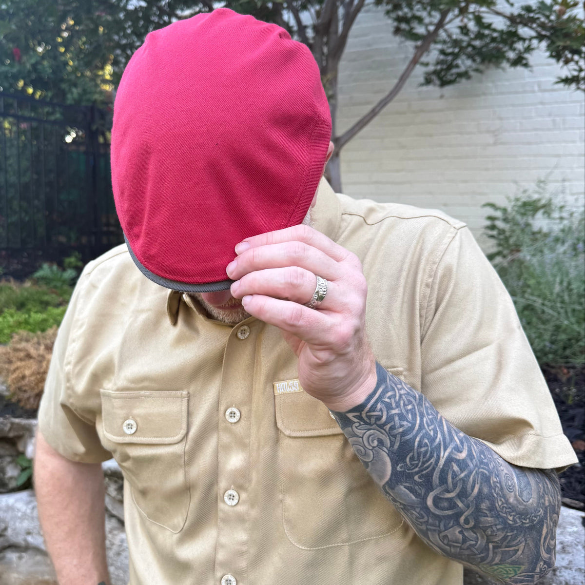 A man wearing a tan short-sleeve button-up shirt and a bright red knit beanie, holding the beanie off his head with one hand. The photo is taken outdoors in a garden setting.