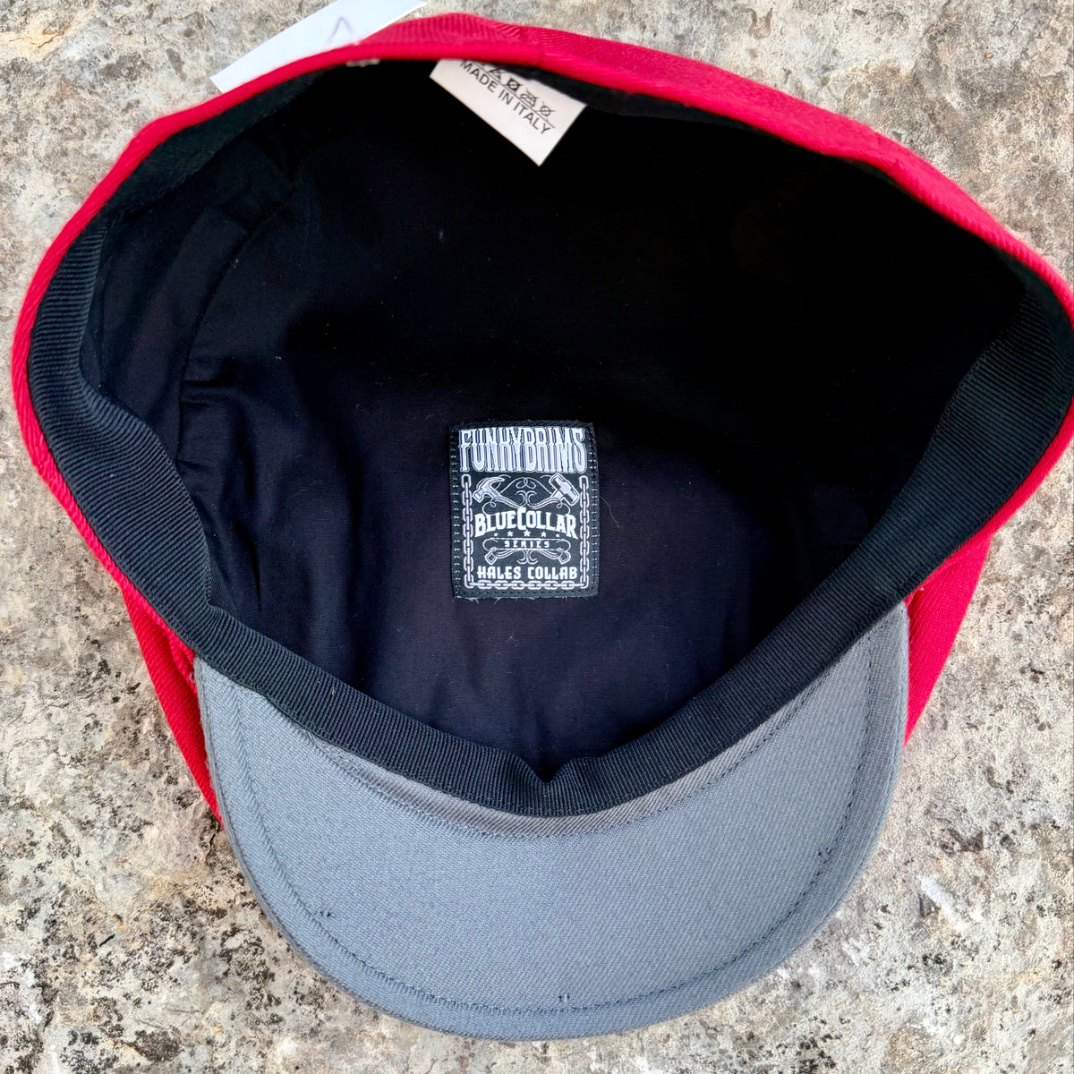A red and gray baseball cap viewed from the inside, showing a black interior lining with a central sewn label that reads 'FUNKYBRIMS BLUE COLLAR 100% HALES COLLAB'. The cap is photographed on a textured stone surface.