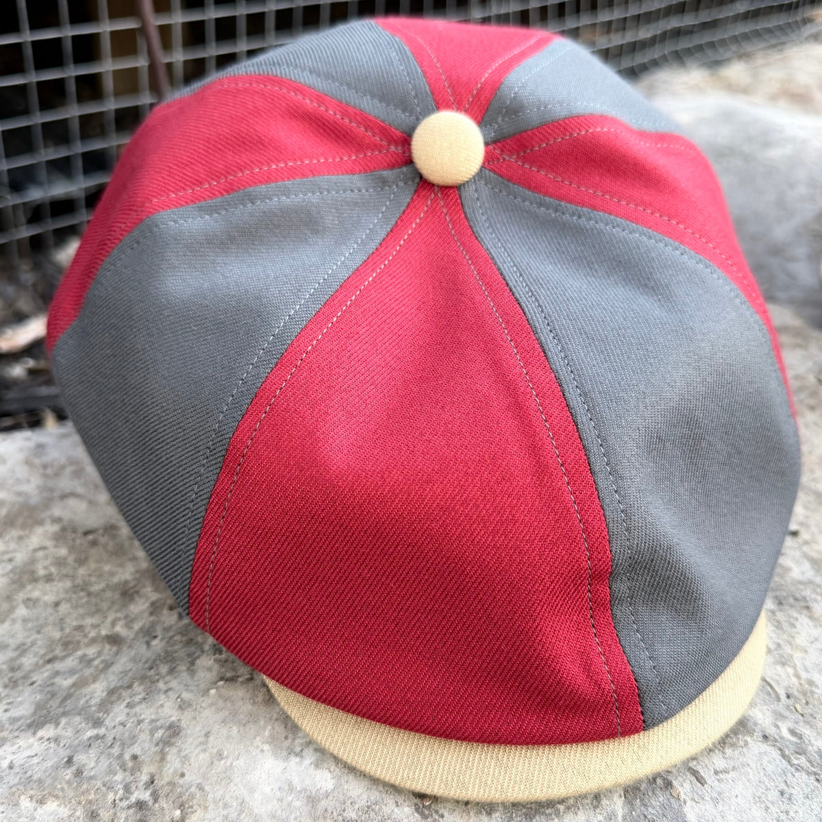 A vintage-style newsboy cap with red and gray panels, a beige button on top, and a beige visor, photographed outdoors on a stone surface.