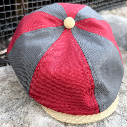 A vintage-style newsboy cap with red and gray panels, a beige button on top, and a beige visor, photographed outdoors on a stone surface.