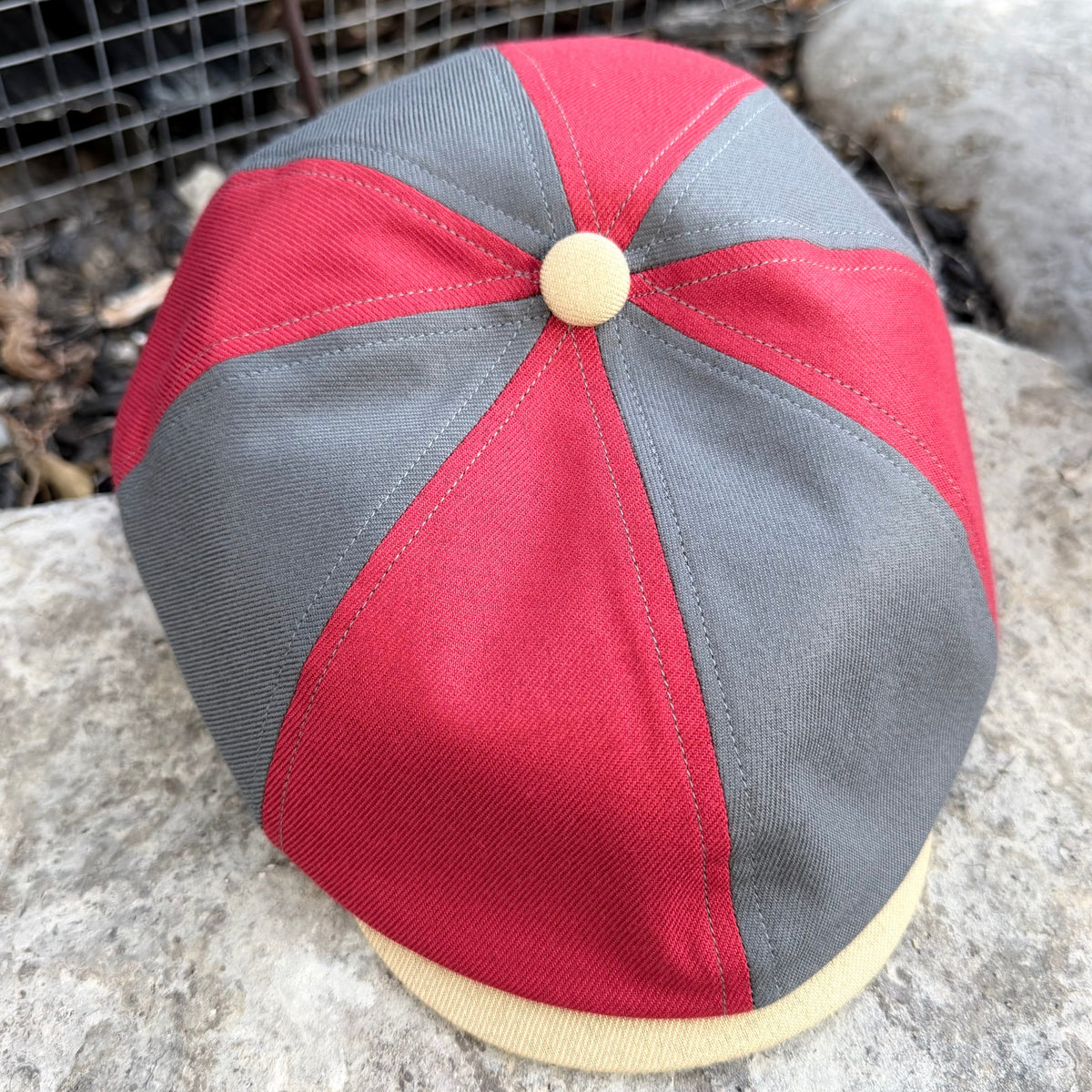 A red and gray newsboy cap with a beige button on top and a beige visor, photographed outdoors on a stone surface.