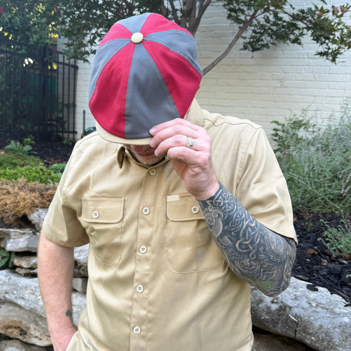A man wearing a beige short-sleeve button-up shirt and a red and gray baseball cap, standing outdoors in front of a stone wall and garden.