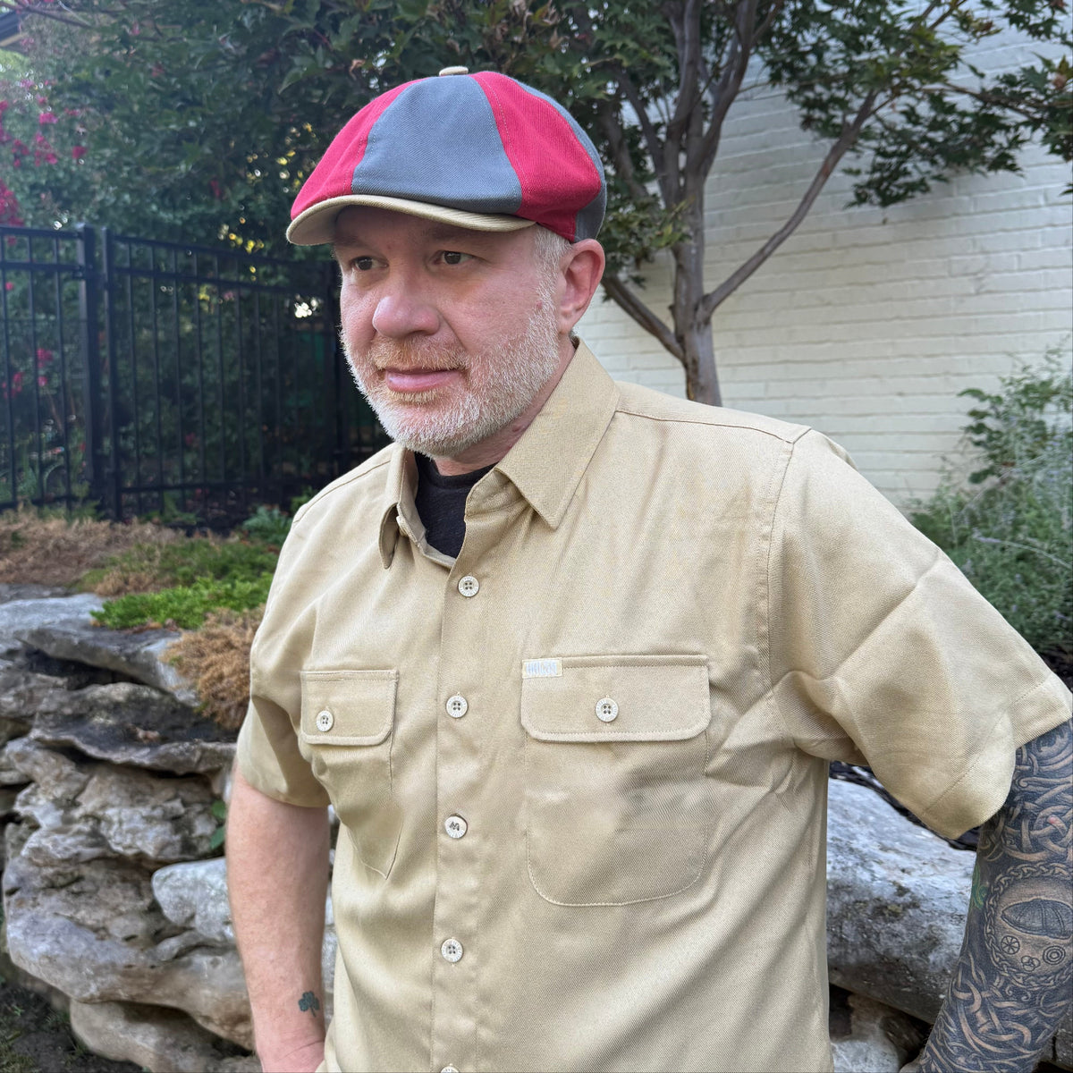 A man wearing a beige short-sleeve button-up shirt and a red and gray flat cap, standing outdoors in a garden setting with stone walls and trees.