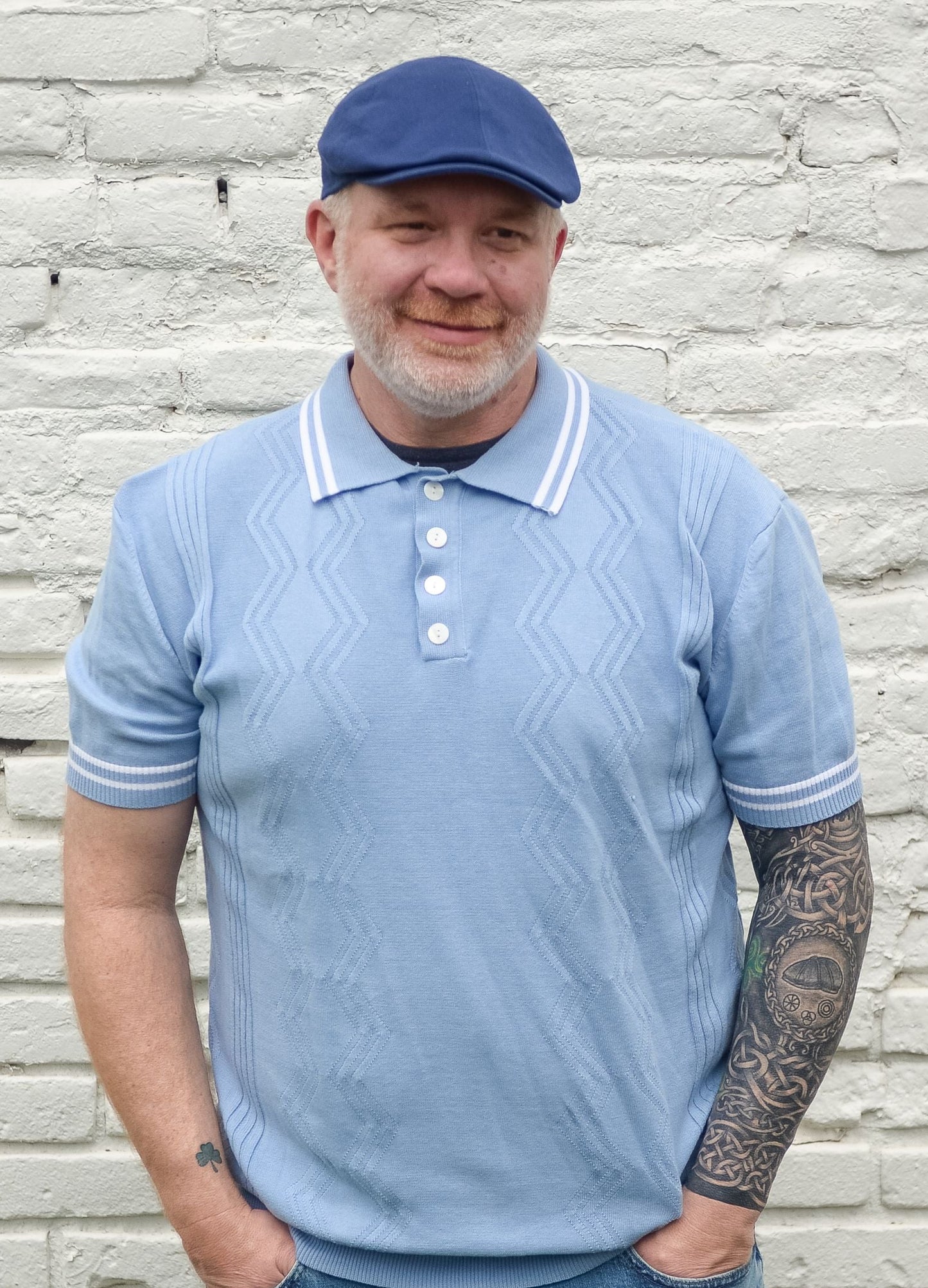 Man wearing a light blue polo shirt and cap standing against a light stone wall.