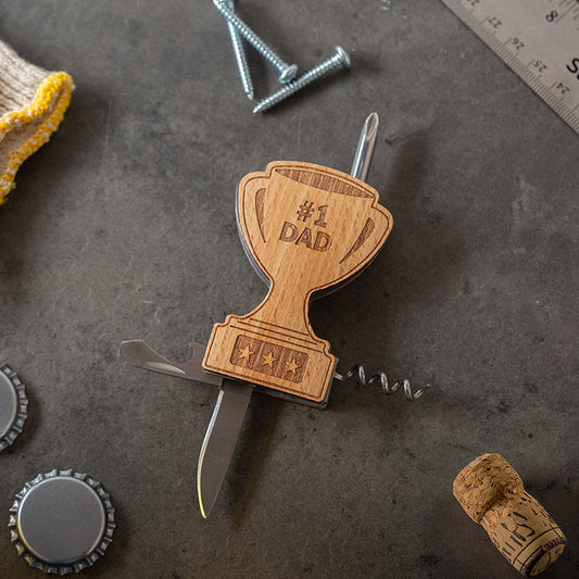 Wooden trophy-shaped multi-tool with '#1 DAD' engraving on a textured surface with tools and bottle caps.