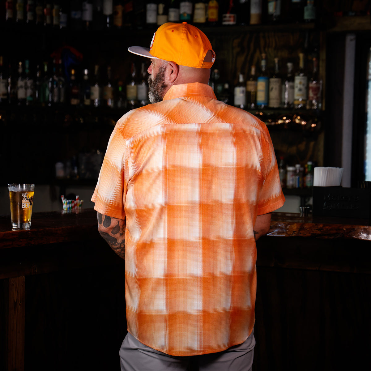 A man standing at a bar, viewed from behind, wearing a bright orange and white plaid short-sleeve button-up shirt and an orange cap. The background shows a dark, out-of-focus bar interior with shelves of bottles.