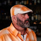 A man with a graying beard wearing an orange and white plaid flat cap and a matching orange plaid button-down shirt, photographed in a dimly lit bar setting.