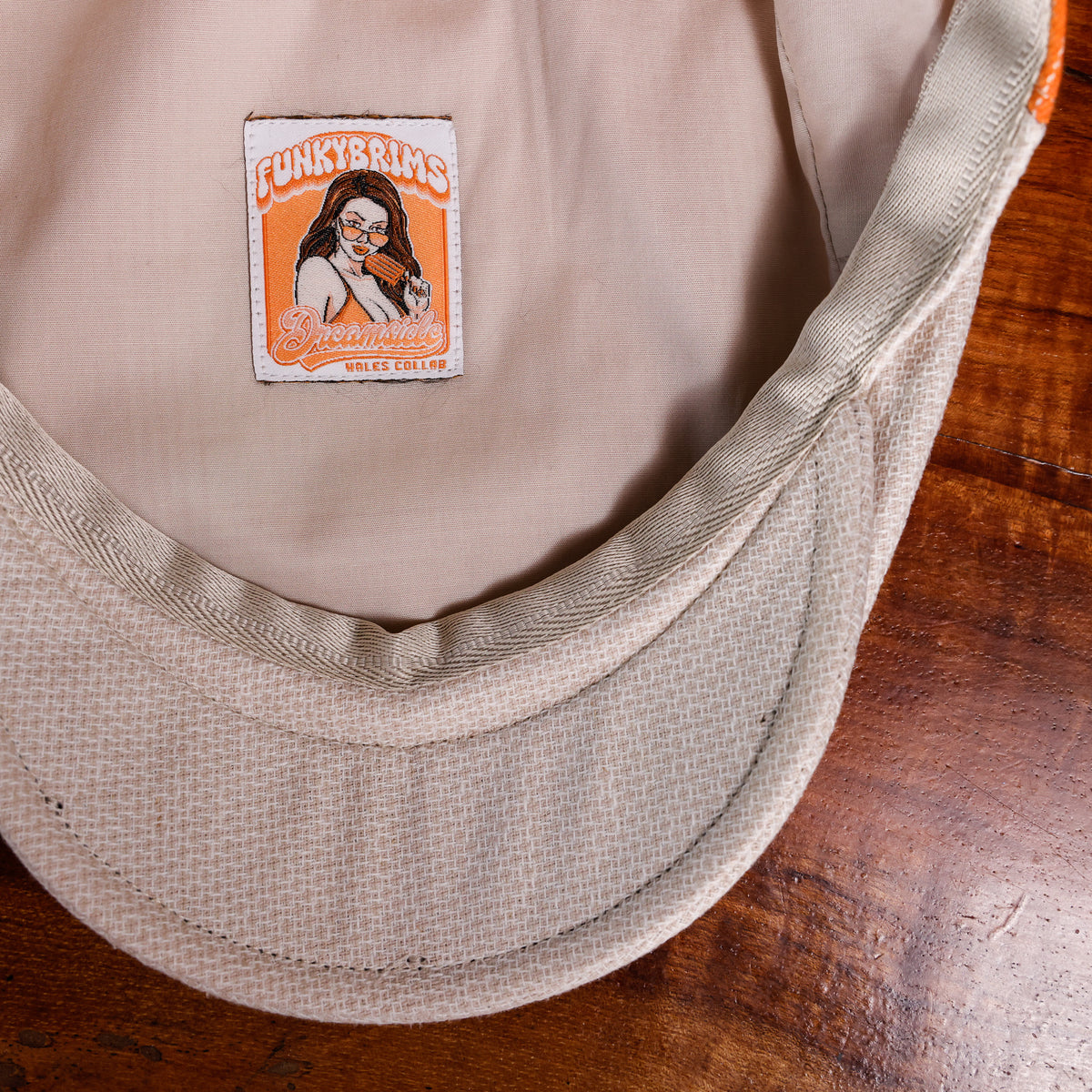 Close-up of the inside of a beige baseball cap resting on a wooden surface, showing a sewn-on patch with an illustration and the text 'FUNKYBRIMS' .