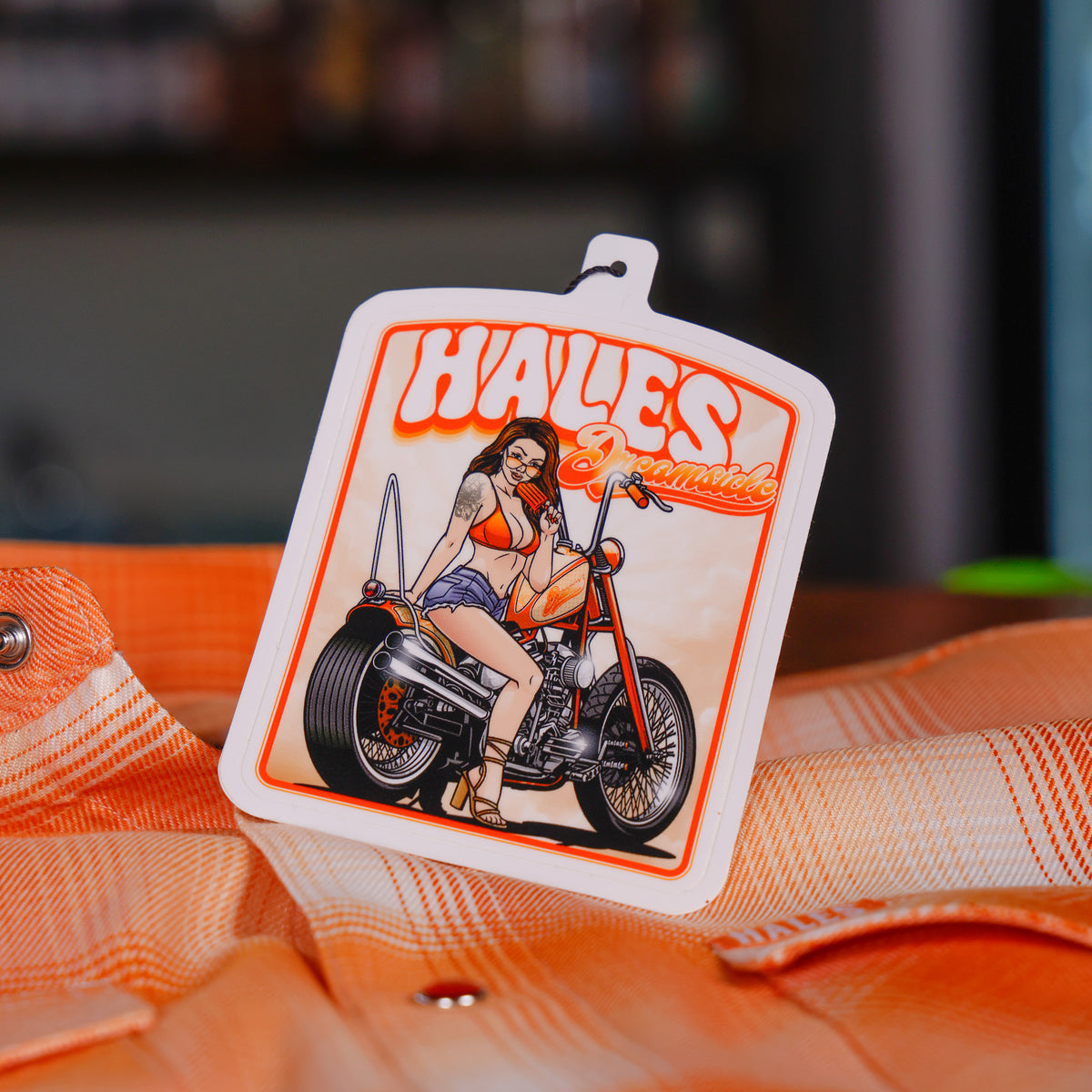 A square white Hales Custom motorcycle-themed keychain or tag featuring a retro illustration of a woman on an orange chopper motorcycle, resting on an orange plaid shirt.