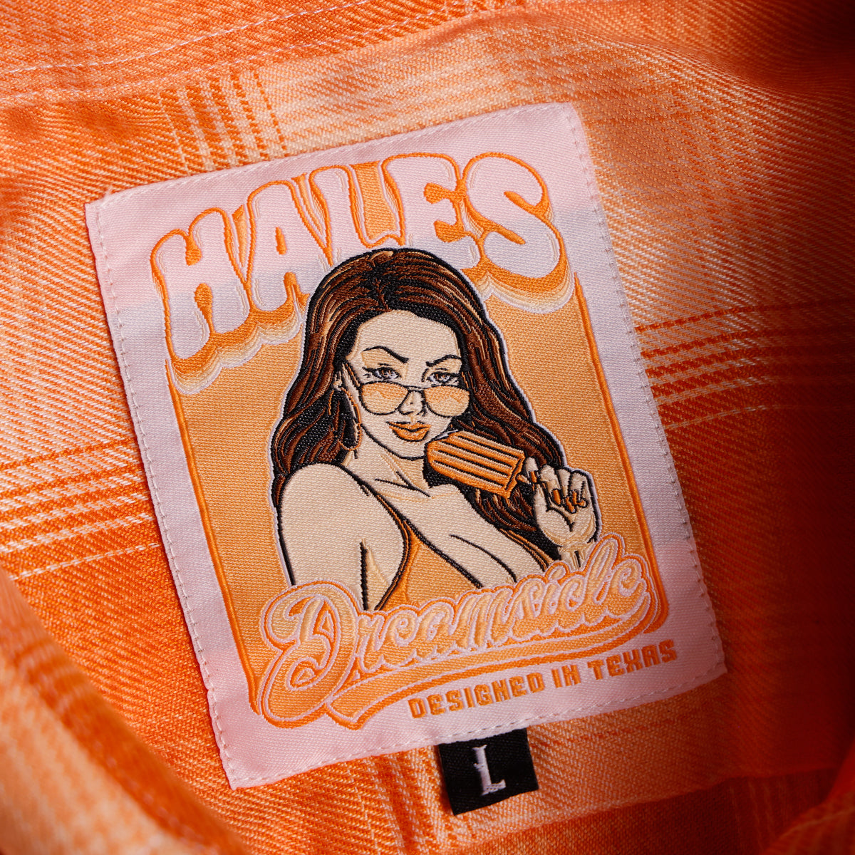 Close-up of an orange plaid garment featuring a sewn-on brand patch with an illustrated woman and the text 'HALES Dreamsicle DESIGNED IN TEXAS', plus a small black size tag marked 'L'.