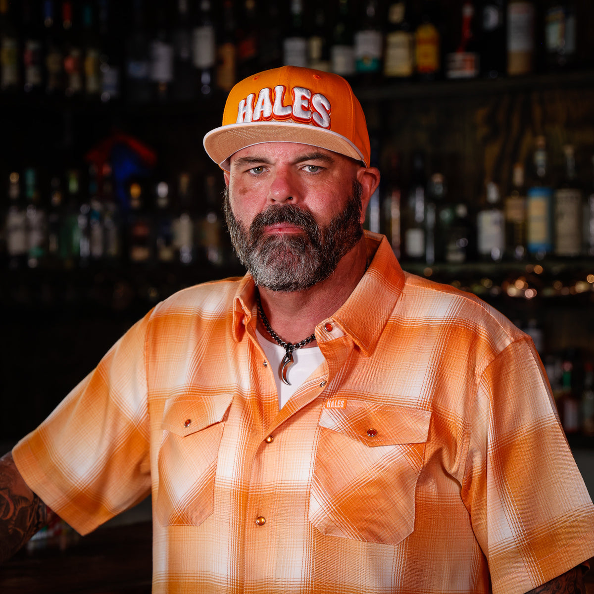 A man with a beard wearing an orange and white plaid button-down shirt and an orange baseball cap with 'HALES' in white letters, standing in front of a dark bar background filled with bottles.