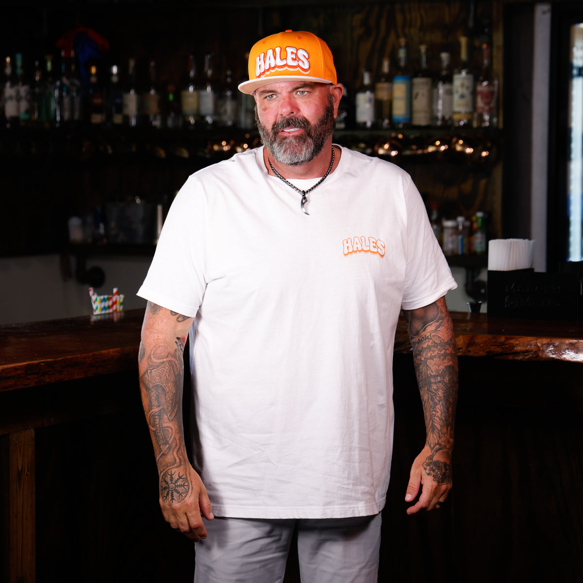 A man with tattoos and a beard stands behind a bar, wearing a white t-shirt and light gray pants, with an orange and white 'HALES' cap on his head. The background shows a dimly lit bar interior with shelves of bottles.