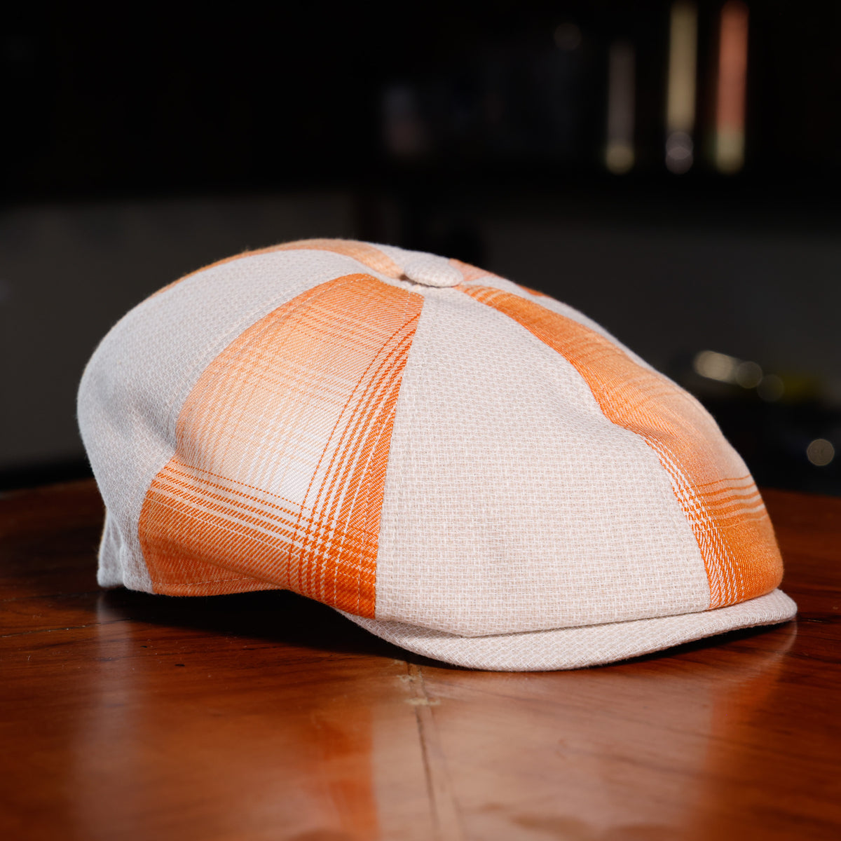 A structured flat cap with a white and orange plaid pattern, resting on a polished wooden table against a dark, blurred background.
