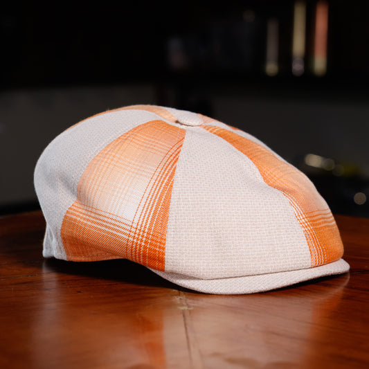 A structured flat cap with a white and orange plaid pattern, resting on a polished wooden table against a dark, blurred background.