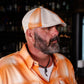 A man wearing an orange and white plaid button-down shirt and a matching orange and white plaid flat-brim cap, photographed in a dimly lit bar setting.