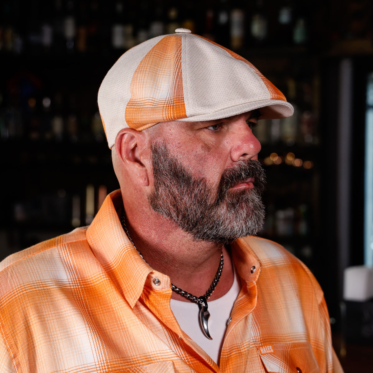 A man wearing an orange and white plaid button-down shirt and a matching orange and white plaid flat-brim cap, photographed in a dimly lit bar setting.