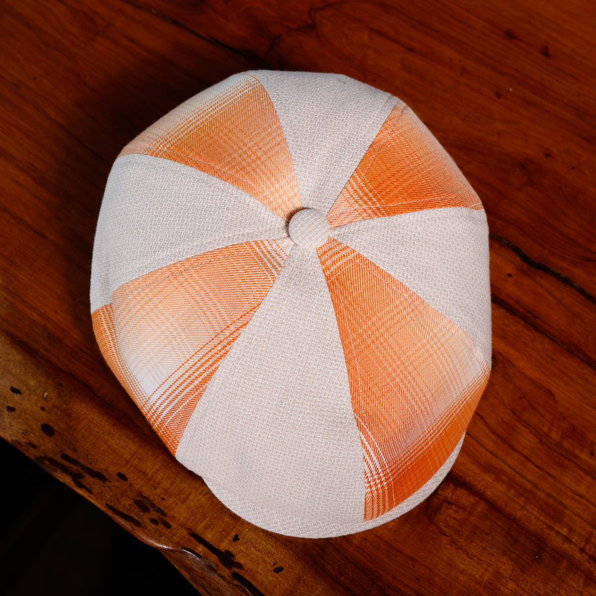 A structured flat cap with alternating white and orange plaid panels, photographed from directly above on a rustic wooden table.