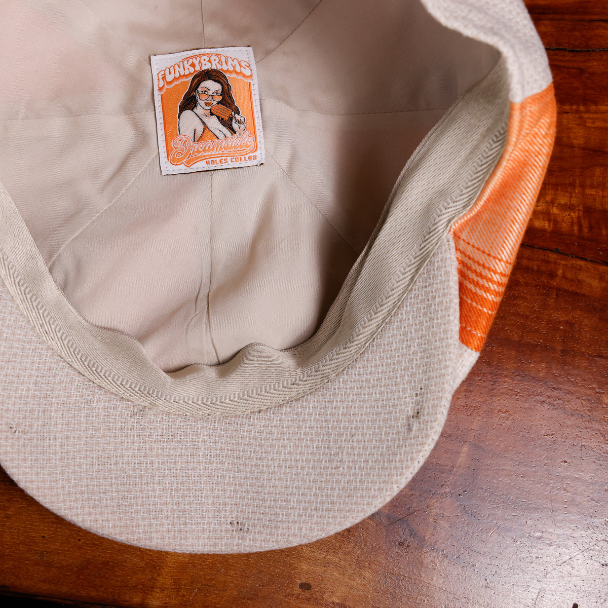 Close-up of the inside of a beige woven cap resting on a wooden surface, showing a sewn-on brand patch that reads Funky Brains and an orange fabric accent on the side.