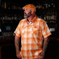 A man with a beard and tattoos wearing a short-sleeve orange and white plaid button-down shirt and a matching orange cap, standing in a dimly lit bar with shelves of bottles in the background.
