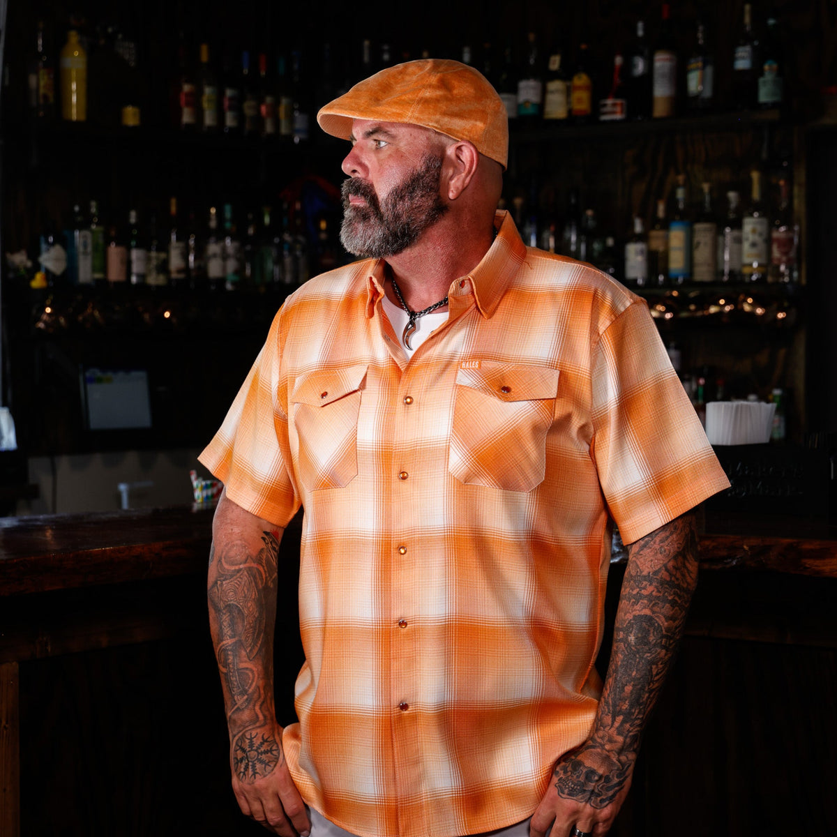 A man with a beard and tattoos wearing a short-sleeve orange and white plaid button-down shirt and a matching orange cap, standing in a dimly lit bar with shelves of bottles in the background.