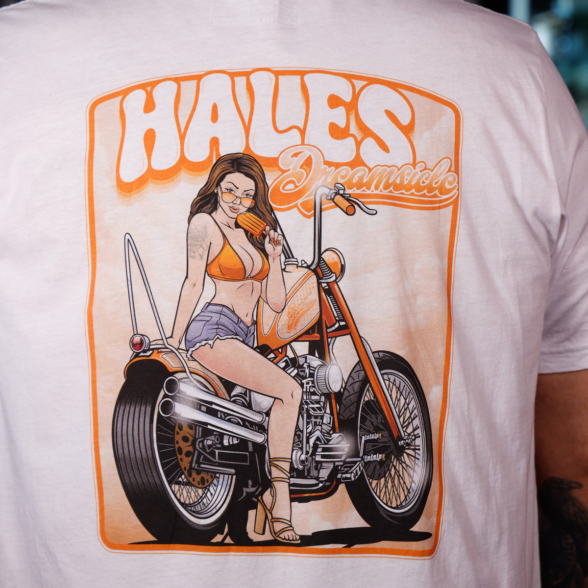 A close-up of a person wearing a white graphic t-shirt. The shirt features a large orange and black illustration of a woman sitting on a chopper motorcycle with the text 'HALES Dreamside' printed above her.