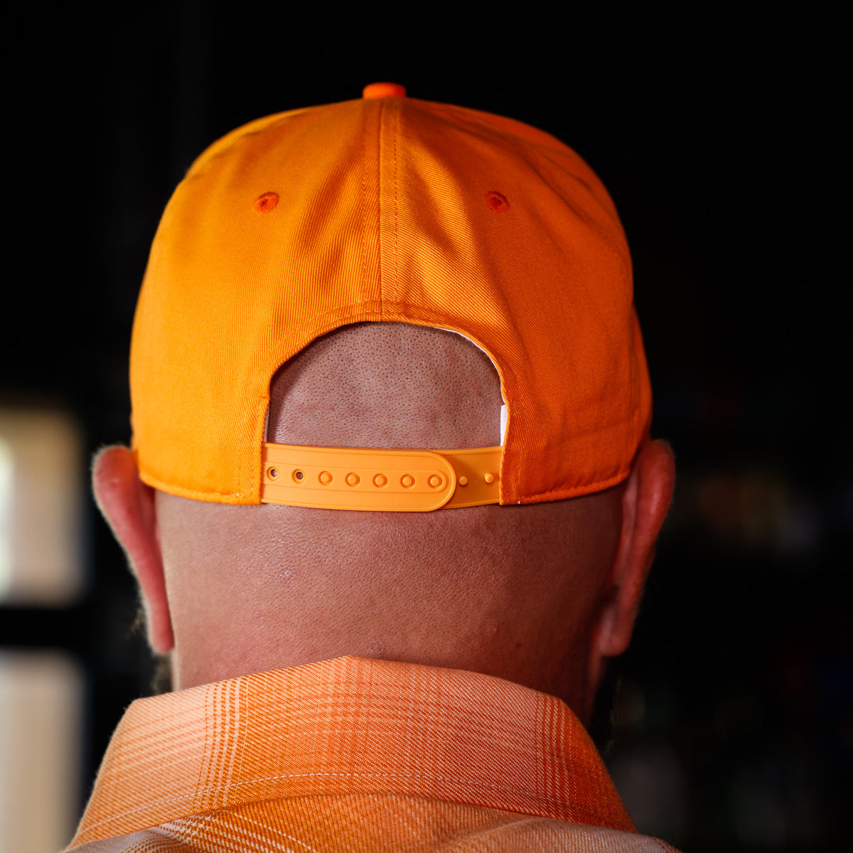 Close-up back view of a man wearing a bright orange baseball cap and matching orange plaid shirt against a dark blurred background.