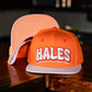 An orange snapback cap with 'HALES' embroidered in white with red and black outlines on the front, displayed on a dark wooden table with another cap behind it.