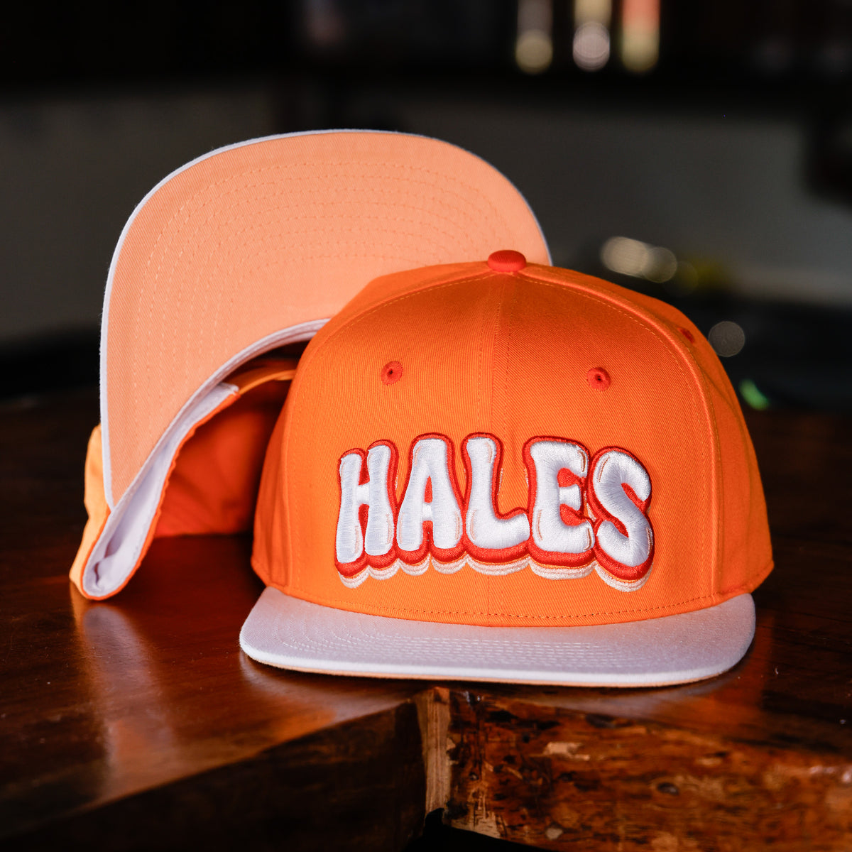 An orange snapback cap with 'HALES' embroidered in white with red and black outlines on the front, displayed on a dark wooden table with another cap behind it.