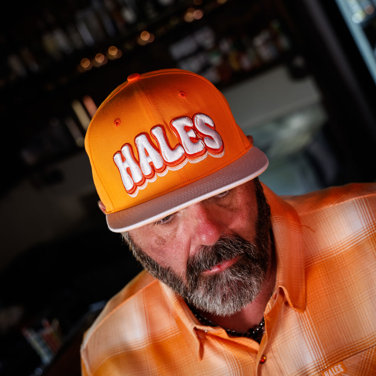 A close-up lifestyle shot of a man wearing an orange snapback cap with 'HALES' embroidered in white with a red outline on the front, paired with an orange plaid button-down shirt.
