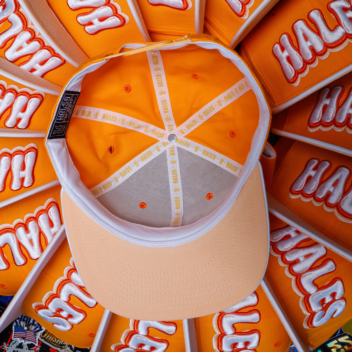 An orange baseball cap shown from above, with a beige visor and a white crossbar. The inside of the cap features orange panels with white and red 'HALES' text, and a black brand tag on the side.