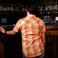 A woman viewed from behind wearing a short-sleeve orange and white plaid button-up shirt, standing at a wooden bar in a dimly lit bar setting.