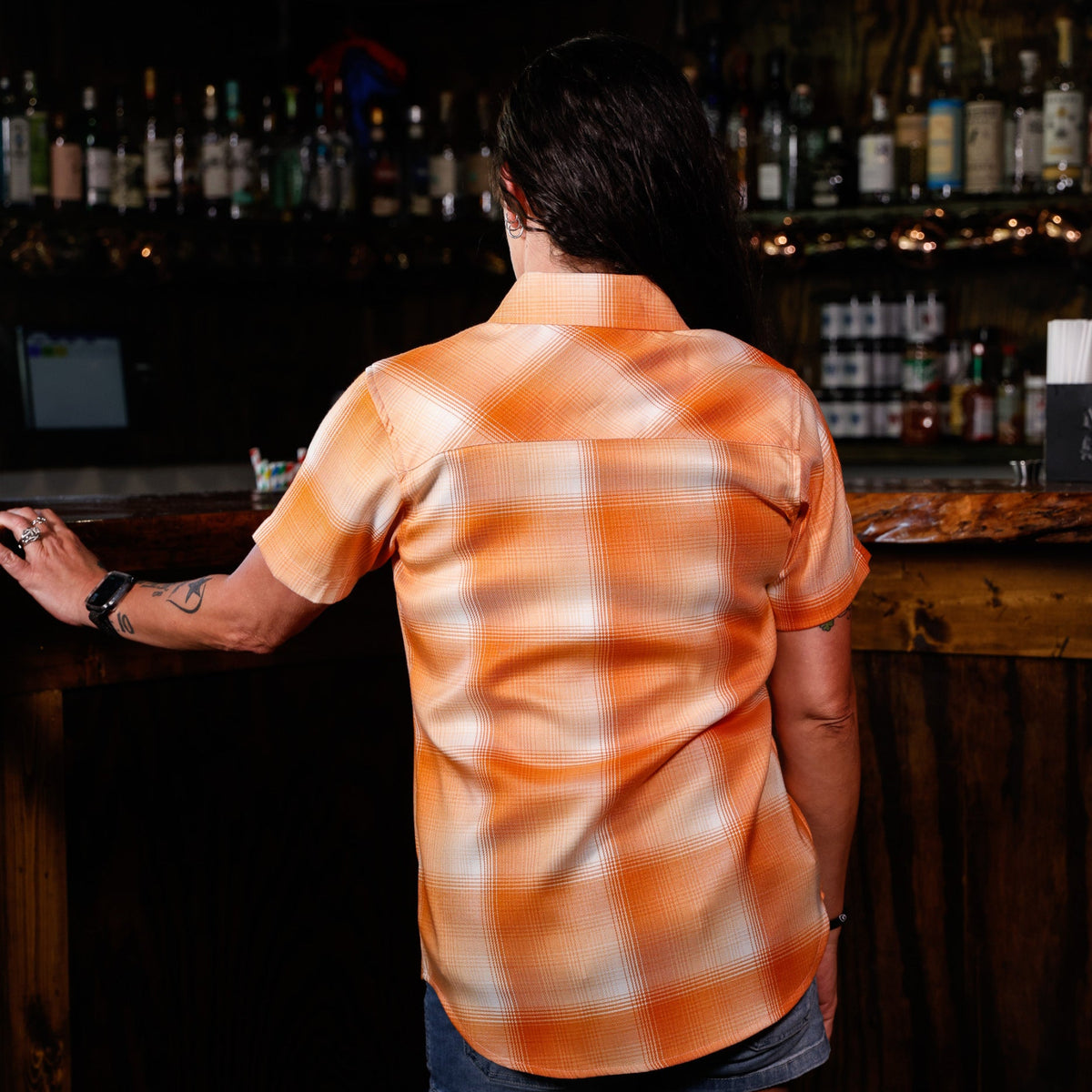 A woman viewed from behind wearing a short-sleeve orange and white plaid button-up shirt, standing at a wooden bar in a dimly lit bar setting.