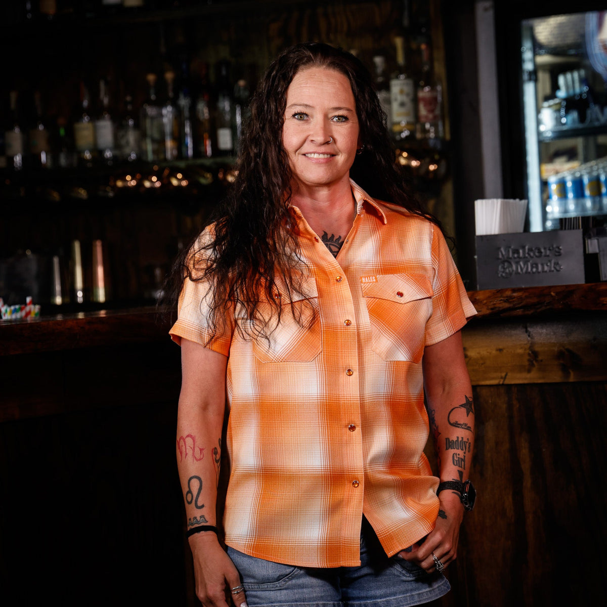 A woman with long dark hair stands in a dimly lit bar setting, wearing a short-sleeve orange and white plaid button-up shirt and denim shorts.