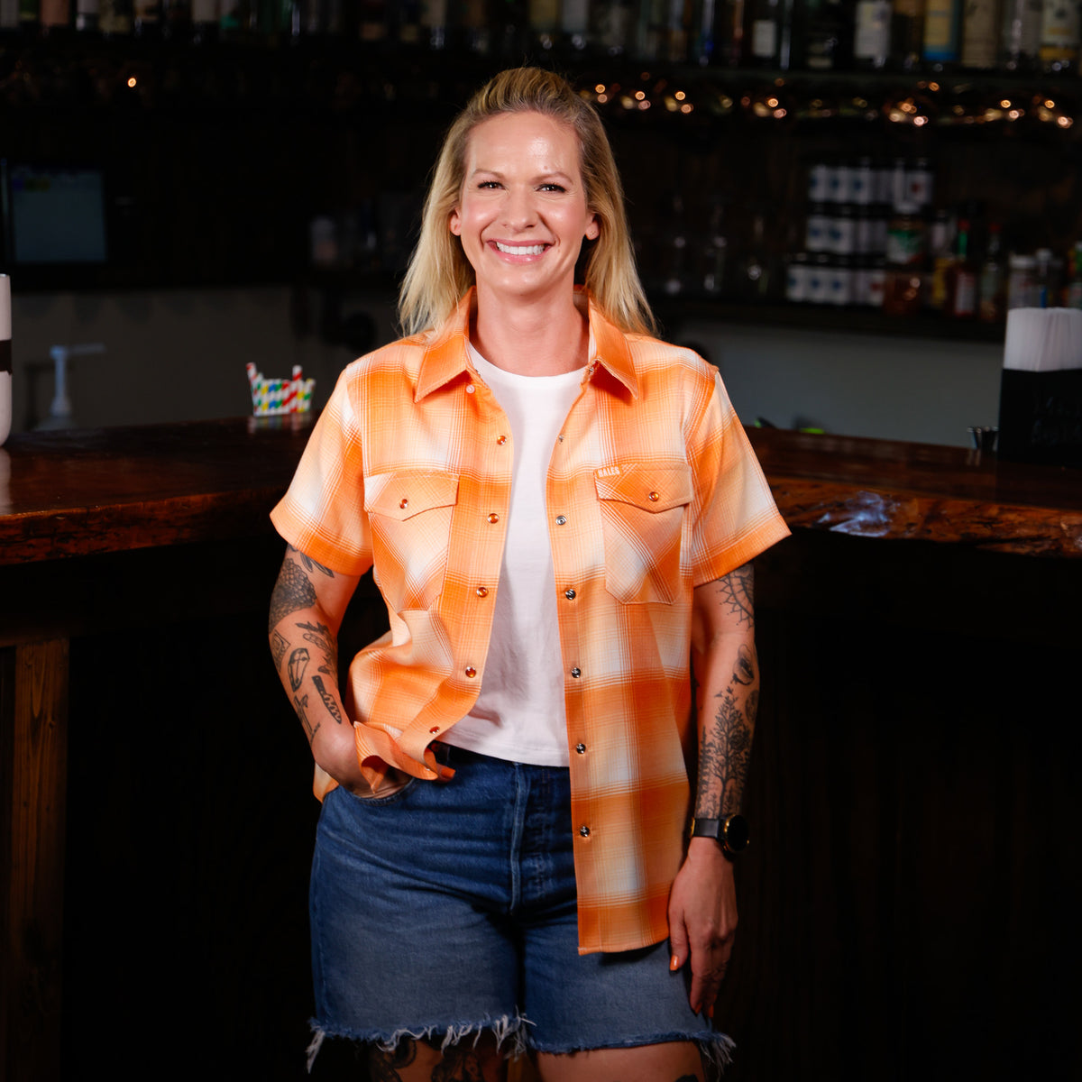 A smiling blonde woman stands in a bar setting wearing a short-sleeve orange and white plaid button-up shirt over a white t-shirt and denim shorts.