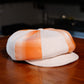A structured flat cap in a cream and orange plaid pattern, resting on a polished wooden table with a blurred indoor background.