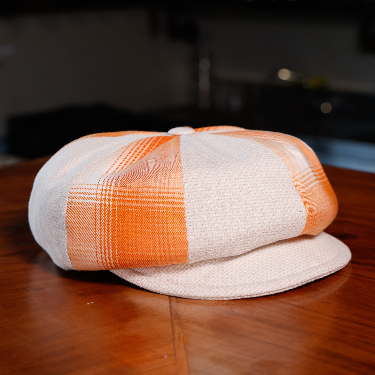 A structured flat cap in a cream and orange plaid pattern, resting on a polished wooden table with a blurred indoor background.