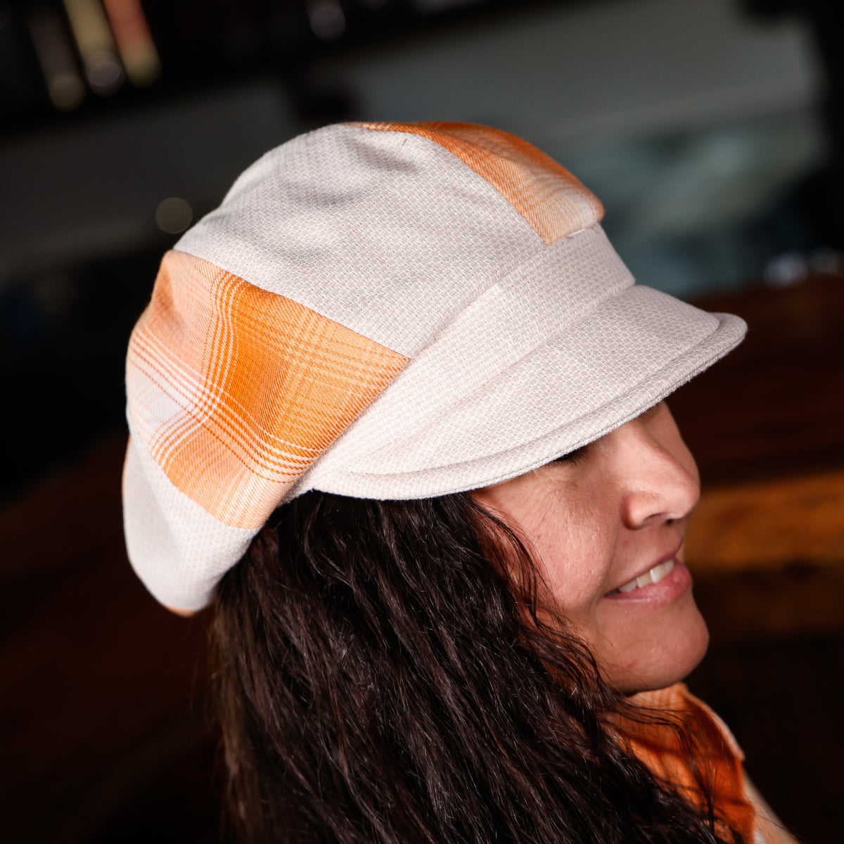 A woman in profile wearing a white and orange plaid sun hat with a brim, photographed in a dimly lit indoor setting.