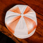 A structured flat cap with a white base and orange plaid stripes, resting on a dark wooden surface.