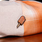 A small enamel pin shaped like a popsicle with orange-red stripes and a tan stick, pinned onto a folded plaid fabric in beige and orange tones, resting on a wooden surface.