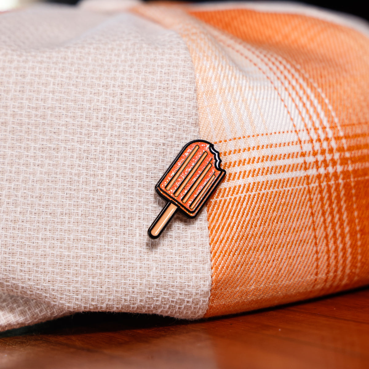 A small enamel pin shaped like a popsicle with orange-red stripes and a tan stick, pinned onto a folded plaid fabric in beige and orange tones, resting on a wooden surface.