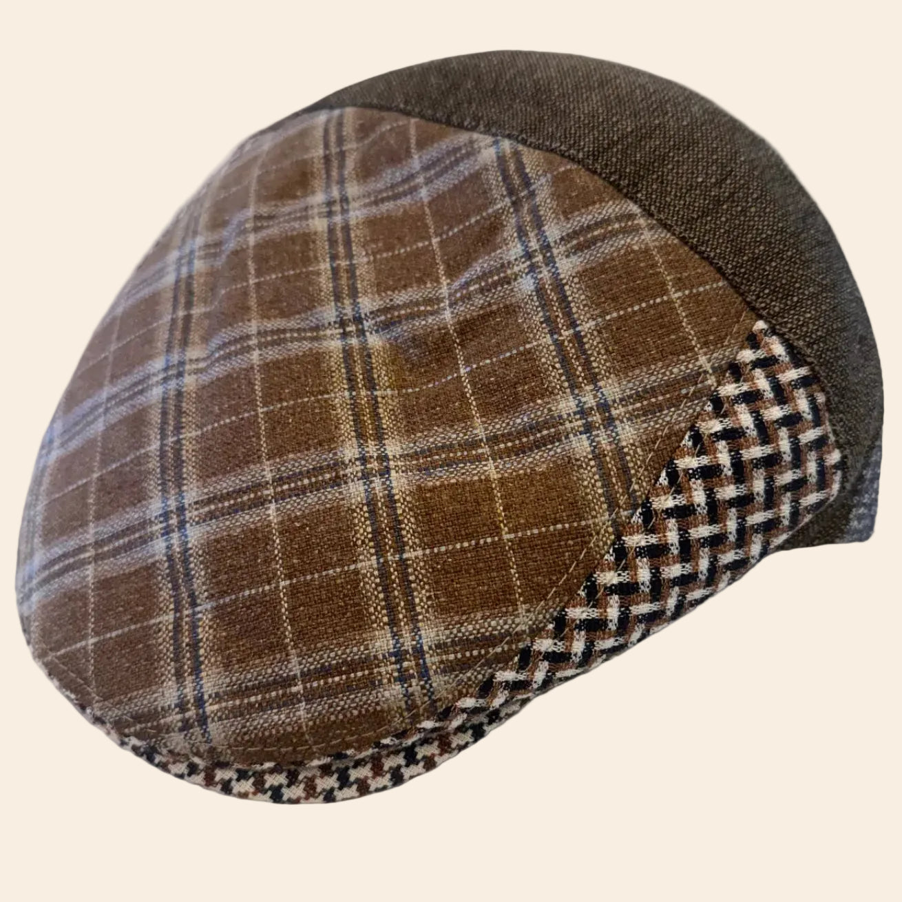A brown plaid flat cap with a dark brown woven side panel and a textured brown fabric crown, resting on a crumpled white surface.